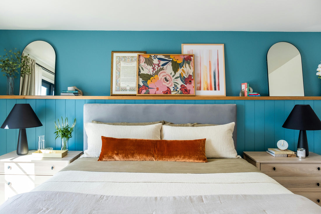 Modern UK bedroom with blue feature wall, wooden shelf ledge above the bed, framed wall art prints, neutral bedding and matching bedside lamps.