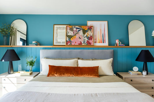 Modern UK bedroom with blue feature wall, wooden shelf ledge above the bed, framed wall art prints, neutral bedding and matching bedside lamps.