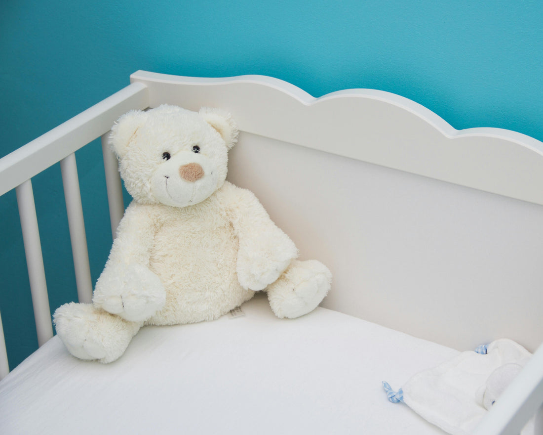 Soft teddy bear sitting inside a white baby cot with a blue nursery wall background in a minimalist baby room decor setting.