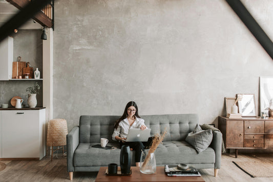 Woman working on a laptop while sitting on a grey sofa in a modern minimalist living room with neutral decor, wooden sideboard and concrete-effect wall