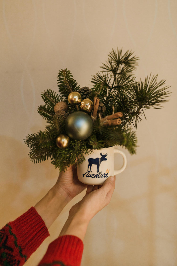 Hands holding a Christmas-themed mug filled with festive greenery, ornaments and pine branches – cozy holiday decor scene.